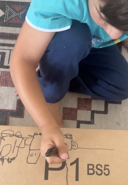 Young child drawing on cardboard as part of a Rooted in Writing hands-on early literacy activity, shared by a parent reviewer from Lakewood, CA.